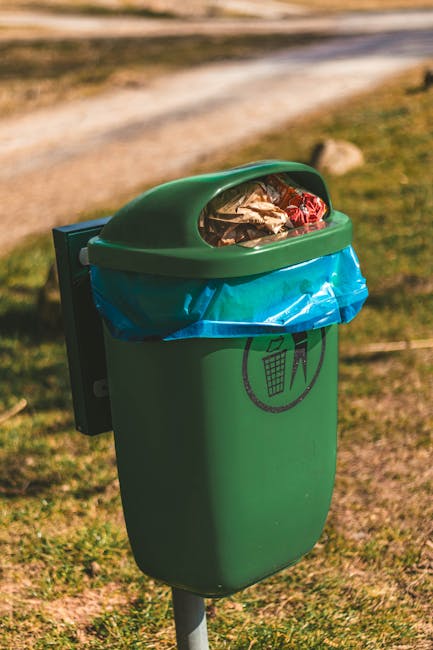 A green outdoor litter bin with a domed, transparent lid filled with various waste items, situated on a grassy area beside a dirt pathway. The bin has a black recycling symbol printed on its front, in