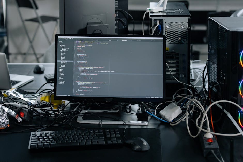 A cluttered workspace featuring a computer monitor displaying lines of code, surrounded by tangled cables and electronic components. To the left, there is a laptop partially covered by aluminum foil, 